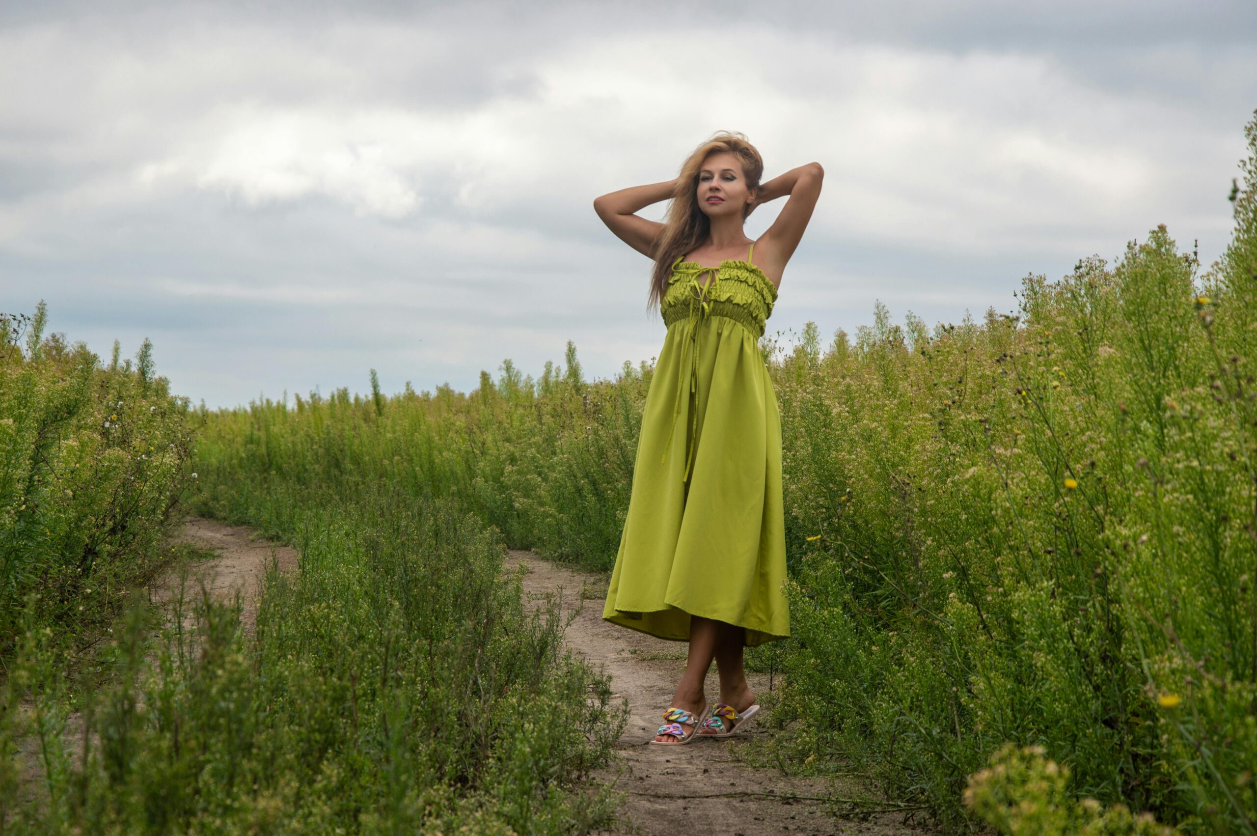 A woman in a green dress standing in a lush field under cloudy skies.